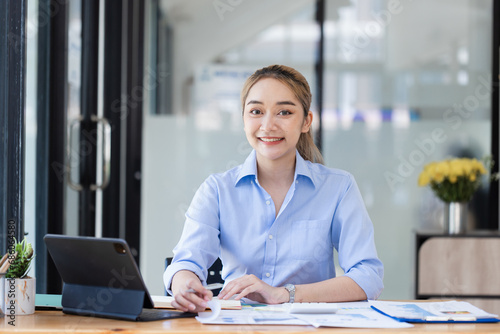 Happy smiling asian businesswoman working on tablet with document paper sitting at desk in office. © SOMKID