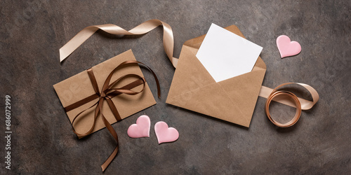 Open brown envelope flat lay with blank card mockup decorated with silk ribbon and silk hearts on dark vintage background. Valentine's Day card. Top view. Banner