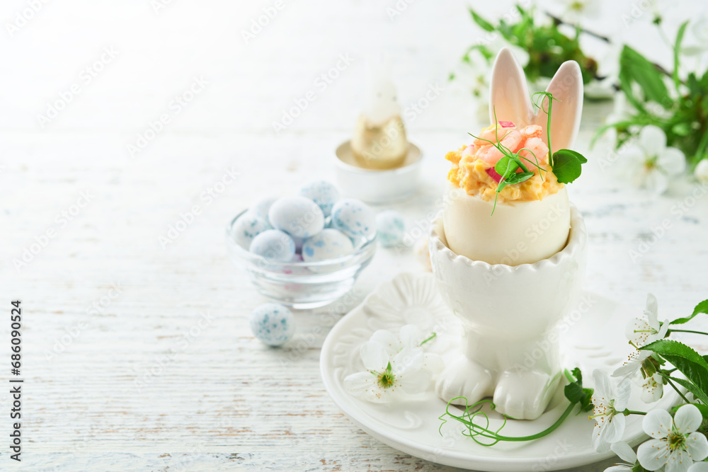 Stuffed or deviled eggs with yolk, shrimp, pea microgreens with paprika in rabbit-shaped stand for easter table decorate fresh cherry or apple blossoms on light background. Traditional dish for Easter Stuffed or deviled eggs with yolk, shrimp, pea microgreens with paprika in rabbit-shaped stand for easter table decorate fresh cherry or apple blossoms on light background. Traditional dish for Easter