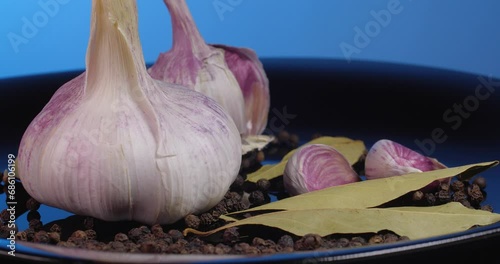 Close up garlic cloves with black peppercorns and  bay leaf rotate on a black plate on a blue background.