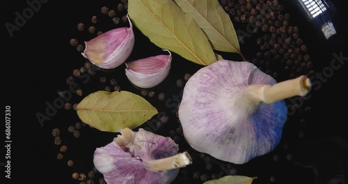 Top view garlic cloves with black peppercorns and  bay leaf rotate on a black plate on a blue background.