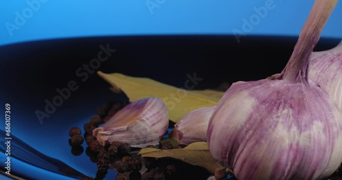 Close up garlic cloves with black peppercorns and  bay leaf rotate on a black plate on a blue background.
