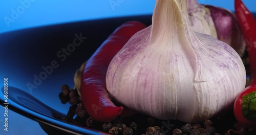 Close up garlic cloves with black peppercorns, red chilly pepper and  bay leaf rotate on a black plate on a blue background.