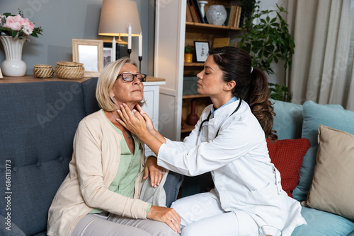 During a home visit to an elderly patient, a young endocrinologist doctor checks her thyroid gland by feeling her neck with her hands. Senior women endocrinology health care photo