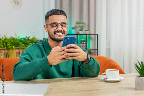 Indian man sits at table uses mobile phone smiles at home office room apartment. Young Arabian guy texting share messages content on smartphone social media applications online watching relax movie