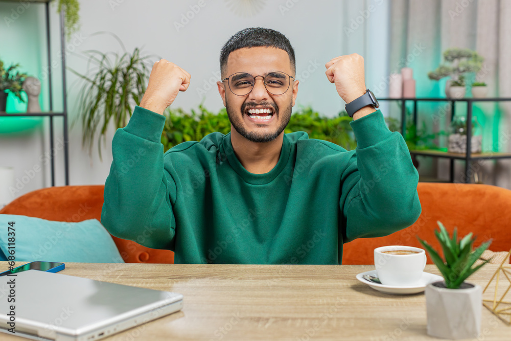 Happy Indian man shouting, celebrating success, winning, goal achievement good news lottery luck victory, watching TV championship at modern home apartment. Arabian guy in office room sits at table Happy Indian man shouting, celebrating success, winning, goal achievement good news lottery luck victory, watching TV championship at modern home apartment. Arabian guy in office room sits at table
