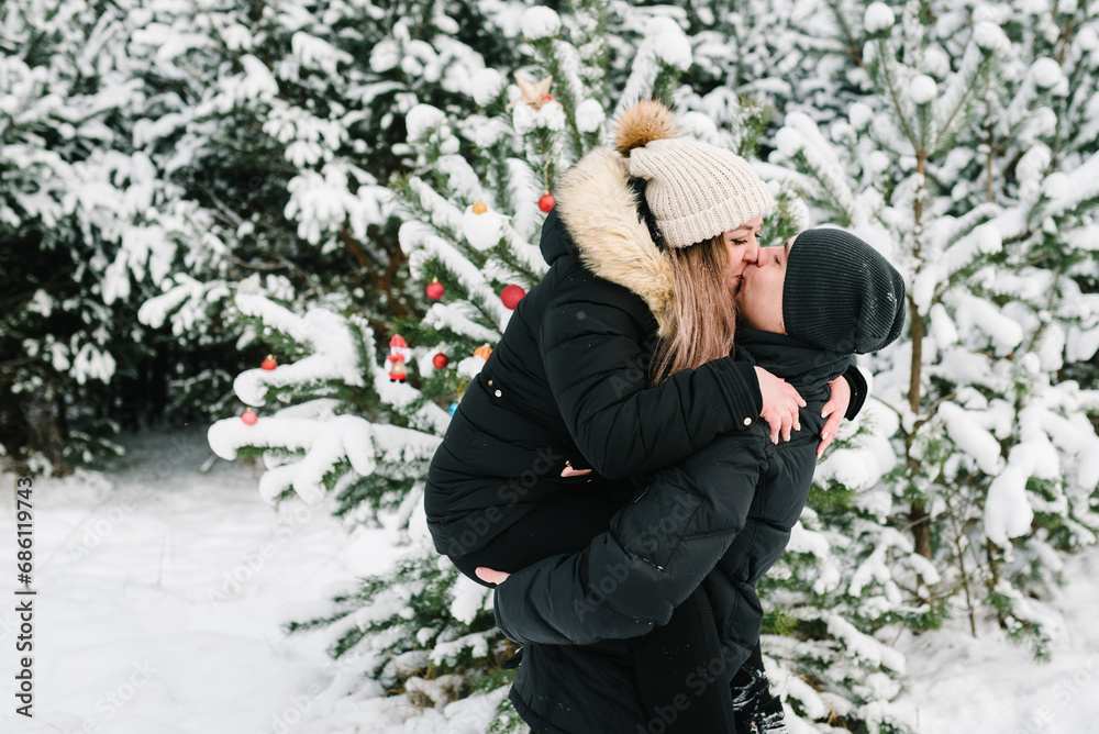 Couple kisses and hugging in snowy winter forest. Young guy and girl in winter wear enjoying snowfall. Man hugs woman standing near decorated Christmas tree in park. Happy winter holidays.