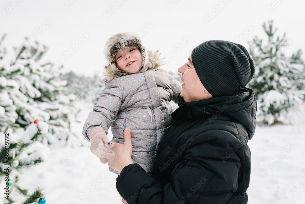 Dad hugging daughter standing in snow in winter forest. Winter holidays. Closeup. Father embracing child girl near Christmas tree in park. Happy family walking snowy mountains, spending time together.