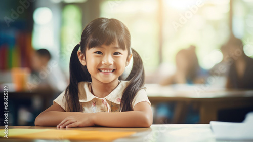 portrait of a smiling asian school girl sitting in front of school desk on a blurred classroom background with classmates photo