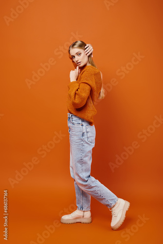 alluring blonde woman in jeans and warm knitted sweater posing on orange backdrop, winter trend