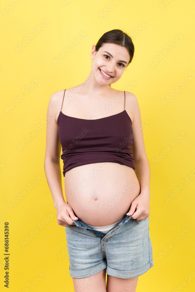 Happy pregnant woman in unzipped jeans showing her naked abdomen at colorful background with copy space Happy pregnant woman in unzipped jeans showing her naked abdomen at colorful background with copy space