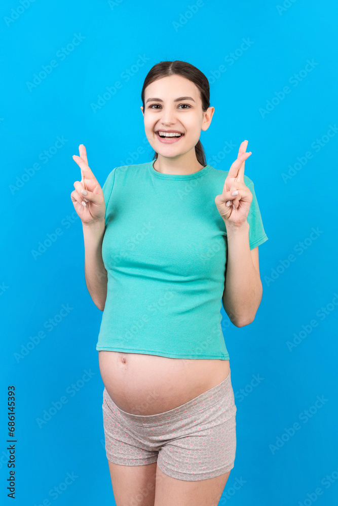 Fototapeta premium Cheerful young pregnant woman holding fingers crossed, waiting pregnancy isolated on colored background in studio. lifestyle pregnancy concept