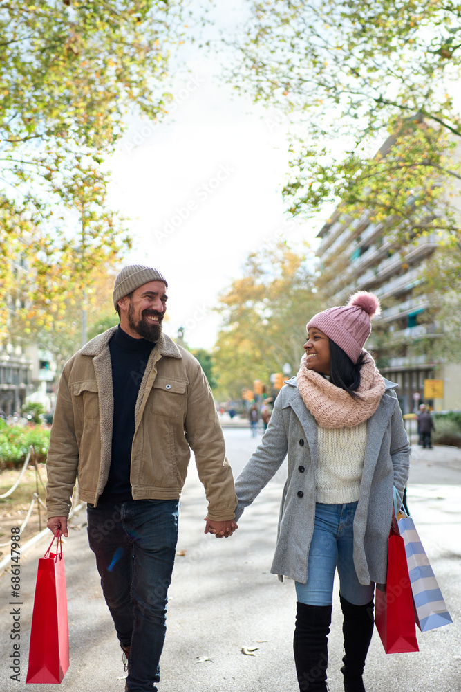 Vertical photo. Beautiful young couple interracial in love enjoying winter holiday season in the city streets, taking a walk, hugging and holding gift bags while celebrating Christmas outdoors