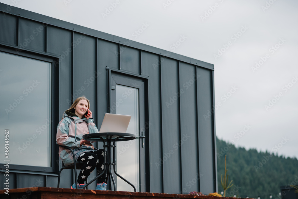 Woman remote work on laptop sitting at table with a great view mountains. Female talking on phone outdoors. Workplace in country in backyard house in morning. Concept freelance lifestyle. Internet 5G.