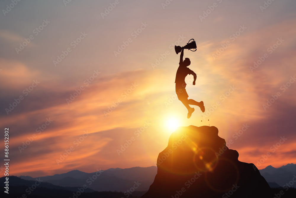 Silhouette of a businessman jumps holding a trophy on top mountain with light sunset. concept of a successful business or determination to lead the organization to success. - obrazy, fototapety, plakaty 