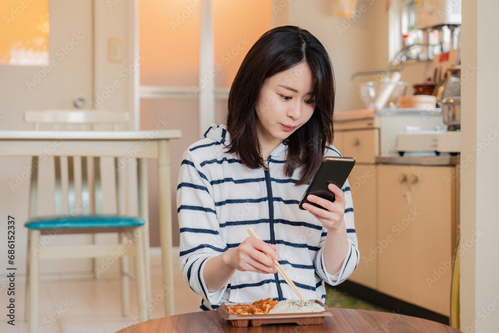 スマホを使いながら家でコンビニ弁当を食べる若い女性 スマホを使いながら家でコンビニ弁当を食べる若い女性