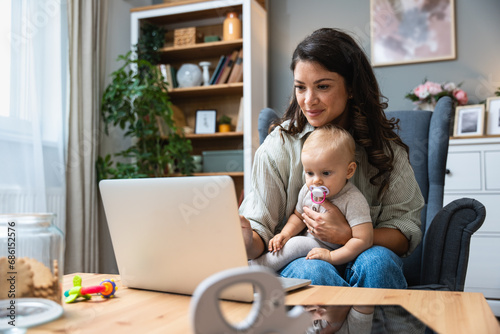 Stay at home mom working remotely on laptop while taking care of her baby. Young business mother on maternity leave trying to freelance by the desk with toddler child. Global work on computer photo