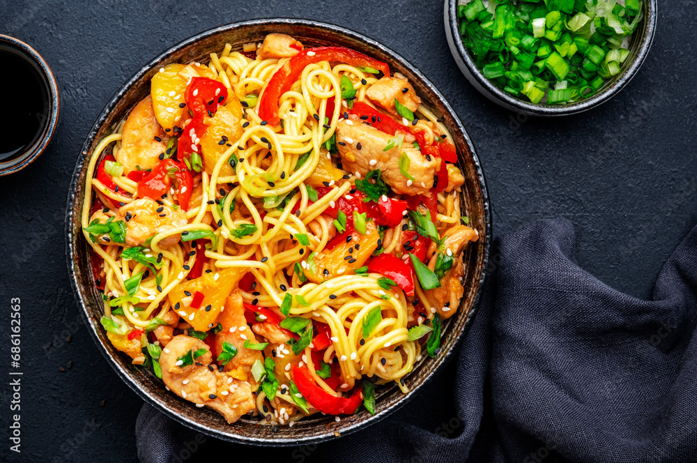 Stir fry noodles with chicken slices, pineapple, red paprika, chives, soy sauce and sesame seeds in ceramic bowl. Asian cuisine dish. Black table background, top view Stir fry noodles with chicken slices, pineapple, red paprika, chives, soy sauce and sesame seeds in ceramic bowl. Asian cuisine dish. Black table background, top view