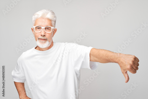 Portrait of elderly gray-haired man showing thumbs down isolated on white studio background. Dislike, wrong choice, negative evaluation concept