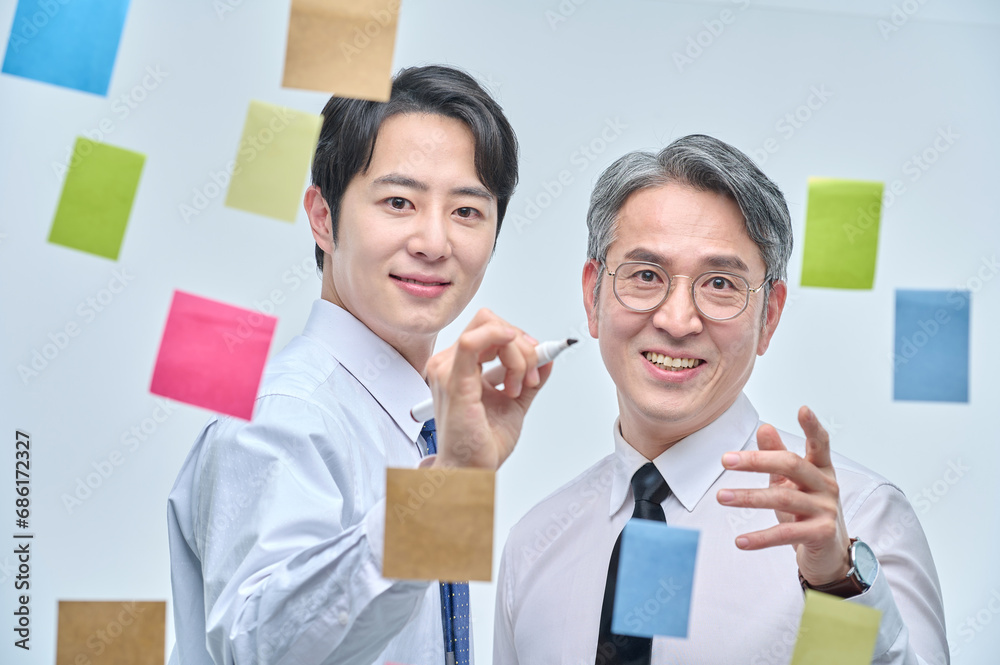 Two businessmen are standing on the glass wall of the office, looking at the attached paper and contemplating planning and strategy. Two businessmen are standing on the glass wall of the office, looking at the attached paper and contemplating planning and strategy.