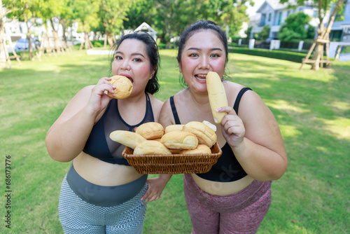 Cheerful two woman overweight in sportswear have fun and enjoy eating bread while exercising in garden. Asian girl chubby holding bread looking at camera standing in the park.