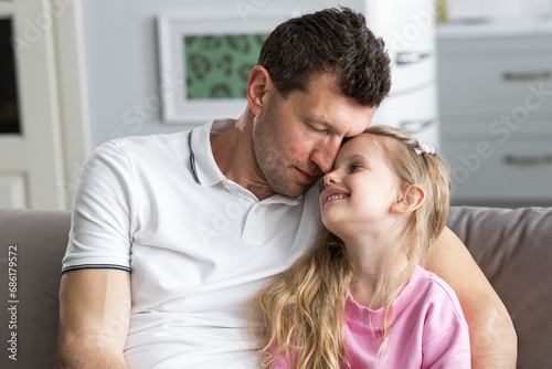 A little daughter sits on the sofa in the room next to her falling asleep father, who hugs her. A girl looks at dad and smiles sweetly. The concept of happy family, childhood and fatherhood. Close up.