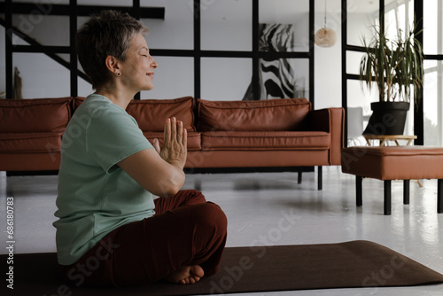 A middle aged lady practices yoga in the large hall. A smiling woman with closed eyes and palms, folded together on chest, sits sideways on a mat on the floor in the lotus position. The concept of photo