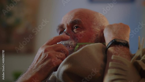 A sick elderly man treats at home independently, puts on an oxygen mask to ease breathing, inhales and exhales, wraps up the plaid on shoulders to keep warm. An ill human suffers from coronavirus or
