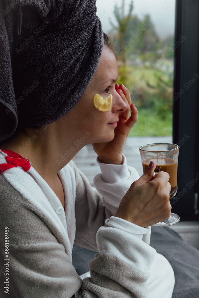 Fototapeta premium Young woman with eye patches and towel drinking morning coffee. Girl after bath with skin mask. Skin care at home. Healthy lifestyle. Pleasure procedure after taking bath. Home cosmetology. 