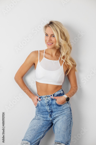 Woman with blonde hair posing near wall. Pretty female with curly hair in white top and jeans.