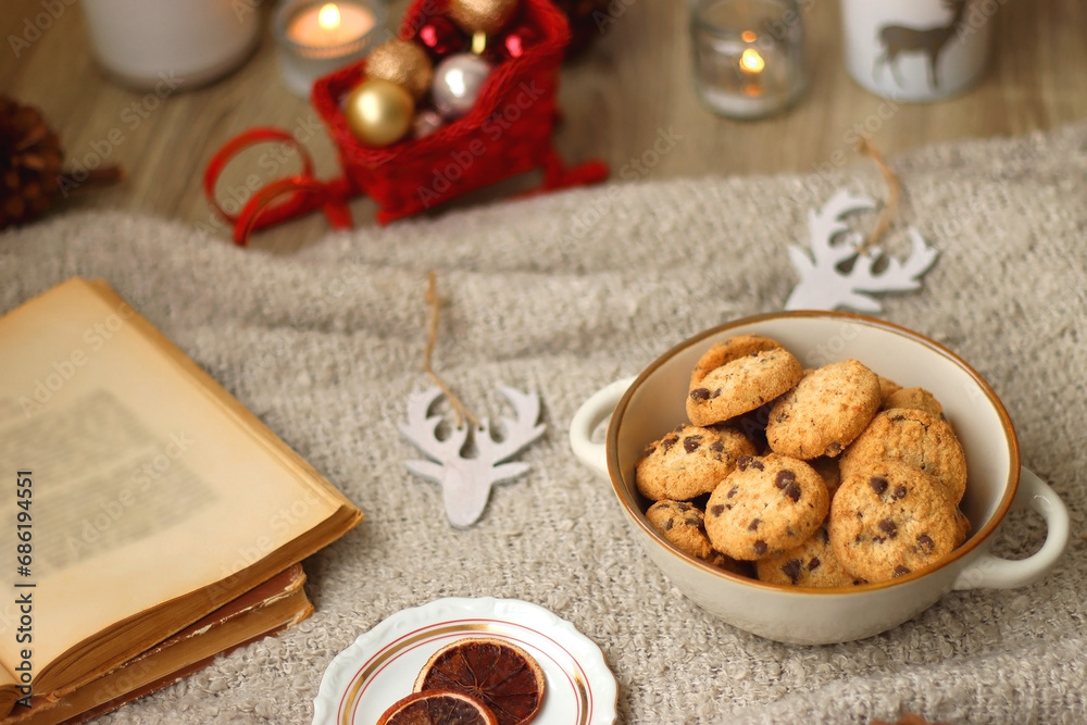 Bowl of cookies, cup of tea, dry oranges, pine cones, books, reading glasses, small presents, various Christmas decorations and lit candles on the table. Cozy Christmas hygge. Selective focus.