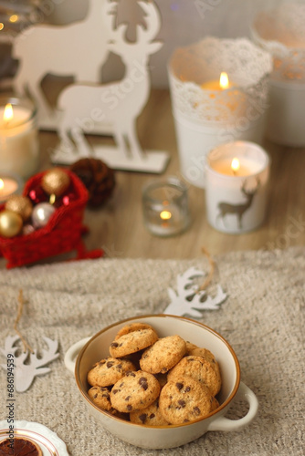 Bowl of cookies, cup of tea, dry oranges, pine cones, books, reading glasses, small presents, various Christmas decorations and lit candles on the table. Cozy Christmas hygge. Selective focus.