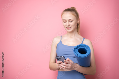 Happy sporty girl with phone and with yoga mat on pink background. Portrait of a slim girl trainer.