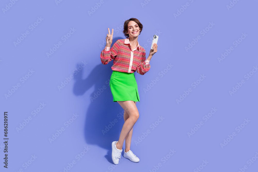 Full size photo of cheerful nice positive woman dressed print shirt holding smartphone showing v-sign isolated on blue color background Full size photo of cheerful nice positive woman dressed print shirt holding smartphone showing v-sign isolated on blue color background