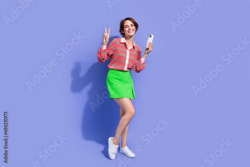 Full size photo of cheerful nice positive woman dressed print shirt holding smartphone showing v-sign isolated on blue color background