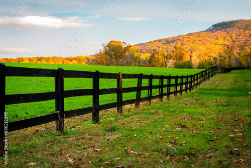 Sunlight shines on Sugarloaf Mountain behind a field of green grass lined by a long wooden fence.