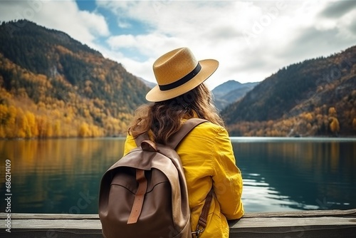 Rear view of a stylish girl, with a backpack, a hat and a yellow jacket, looking at the view of the mountains and the lake in the autumn nature © MaxSimplify