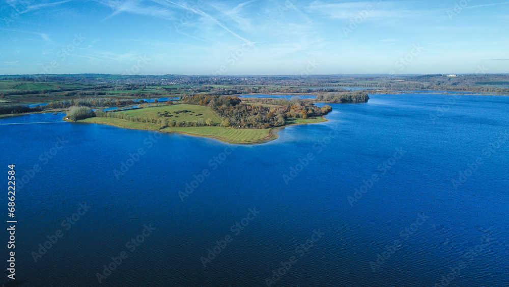 Aerial view 4k wide angle of fields and Rutland water, England, cloudy skies and green patches of forest.