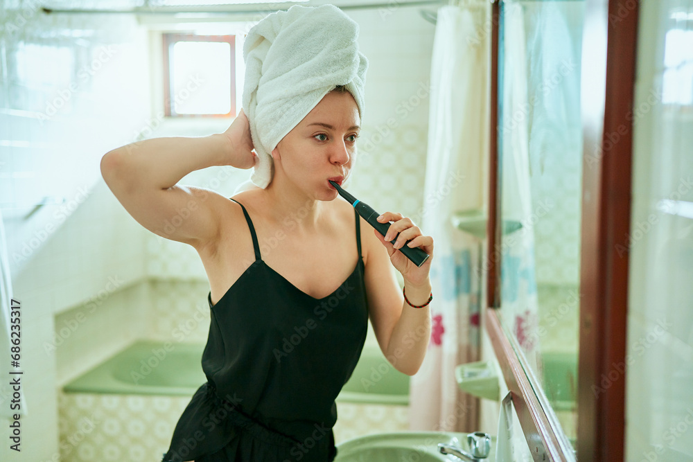 Young adult woman in bathroom with towel on head, promotes dental hygiene using electric toothbrush, mirror reflection shows daily health routine. - obrazy, fototapety, plakaty 