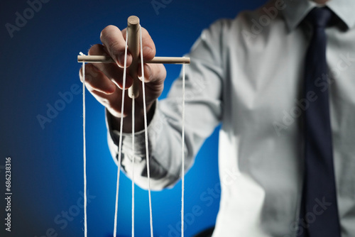In the frame against a blue background stands a man in a suit holding a device with ropes that hold a puppet. Depicts a businessman who manages someone, directs. Theatrical performance