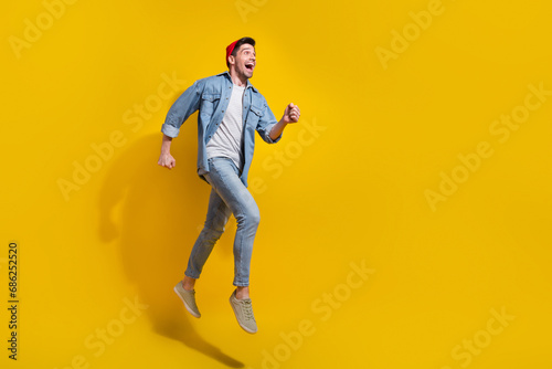 Full length photo of cheerful excited guy wear denim jacket running fast jumping high empty space isolated yellow color background