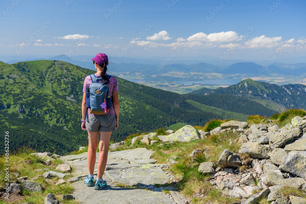 Polish Tatra Mountains, a young woman in shorts and a cap with a mountain backpack stands on a mountain trail and looks at the valley with mountain peaks on a summer sunny day.