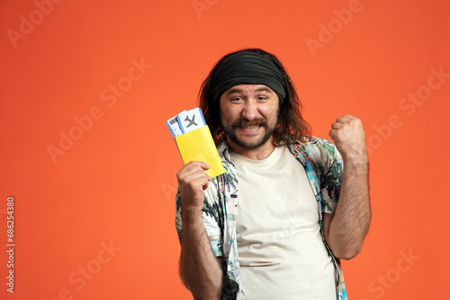 A longhaired man in a bandana holds a passport with airline tickets and makes a victory gesture with his hand. Happy man in studio on orange background close up. photo