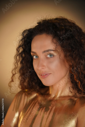 Portrait of female model in studio. Attractive young woman with curly hair and golden makeup with covered chest in golden paint looks at camera.