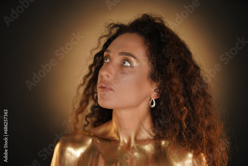 Portrait of female model in studio. Attractive young woman with curly hair and golden makeup looking up, chest coated in golden liquid paint.