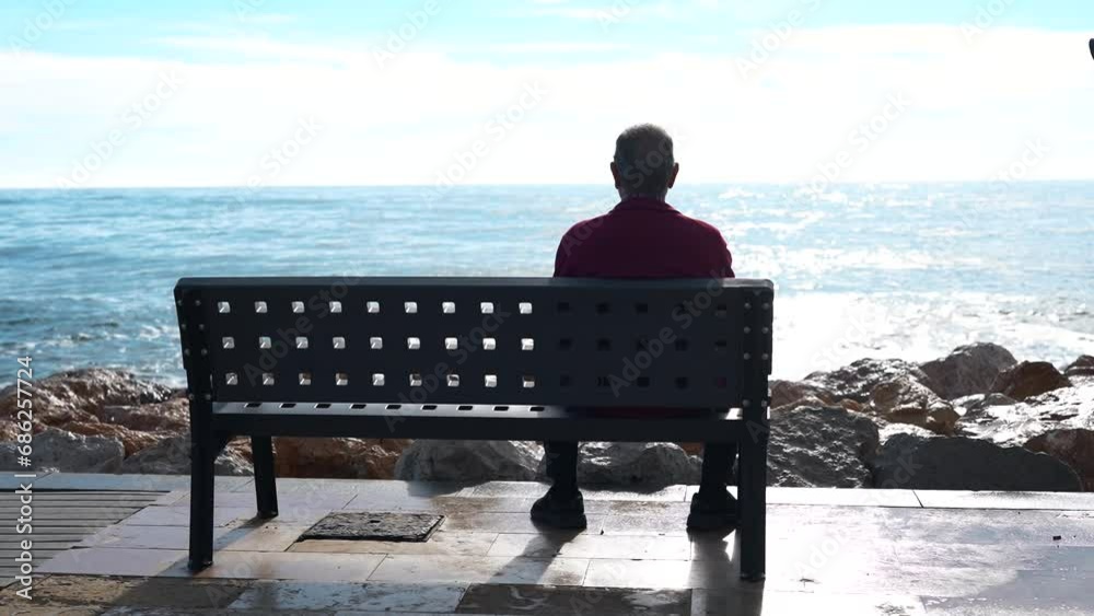 A lonely elderly man sits on a bench and looks at the sea.