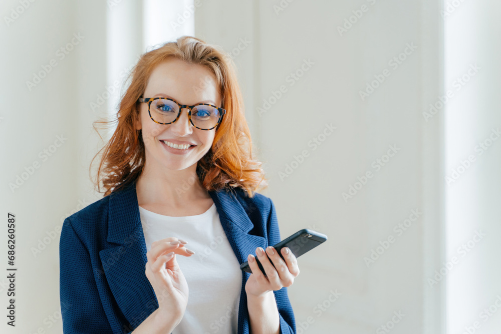 Smiling red-haired woman in blue jacket holding phone, exuding confidence and happiness