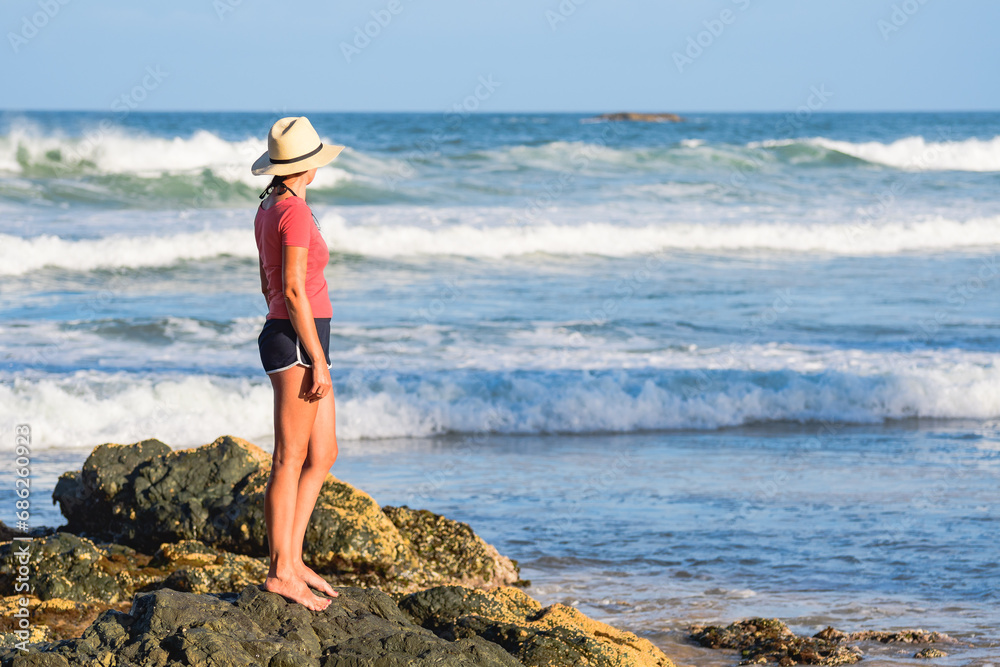Australian coast, a young woman in a hat,T-shirt and shorts stands on the rocks on the beach and looks towards the sea with big waves on a summer sunny day.
