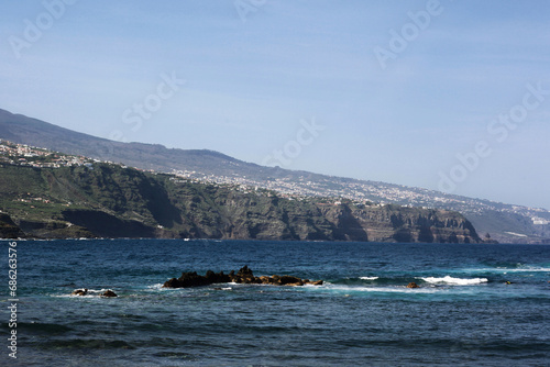 Rocky shore background. Volcanic rock on ocean shore. Tenerife on Canary island landscape. Sea wave crushing. Water white foam. High sea cliff. Turbulent ocean background.