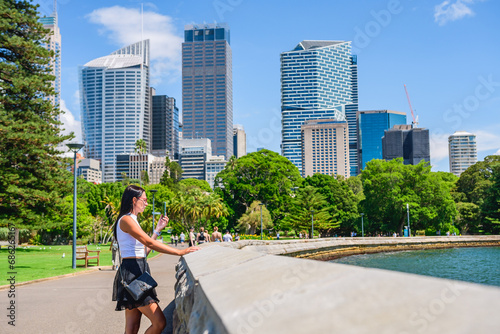 A young woman in a T-shirt and skirt stands in the park on the promenade and talks on the phone, skyscrapers in the business district, view from the Royal Botanic Garden Sydney, summer sunny day.